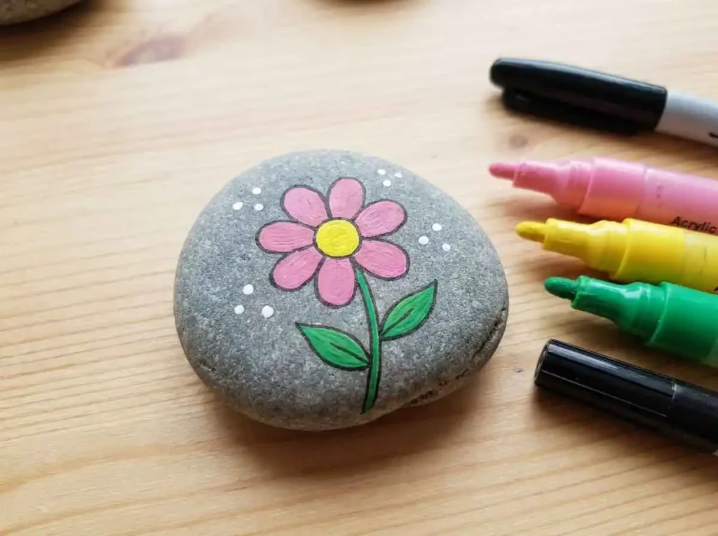 Smooth gray rock decorated with a simple pink flower, yellow center, and green leaves using acrylic paint pens.